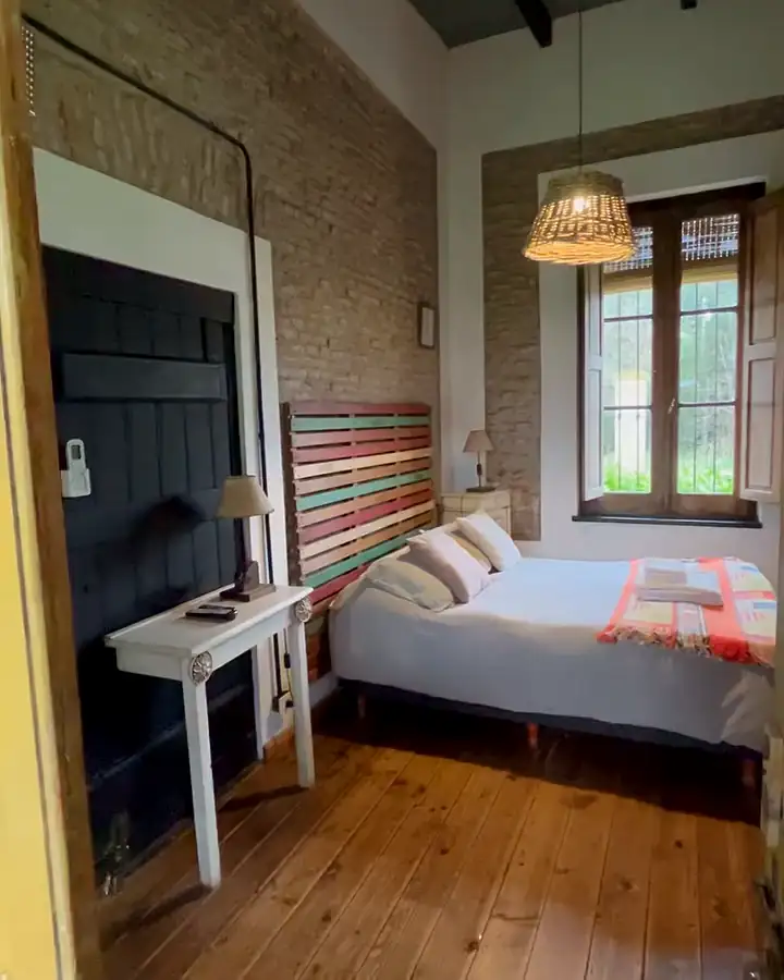 La habitación 8: cama con manta tejida a rayas, pared de ladrillo y una ventana de madera que da al parque.