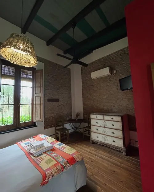 La suite desde otro ángulo: ventana de madera con postigos, lámpara de mimbre y cómoda antigua.